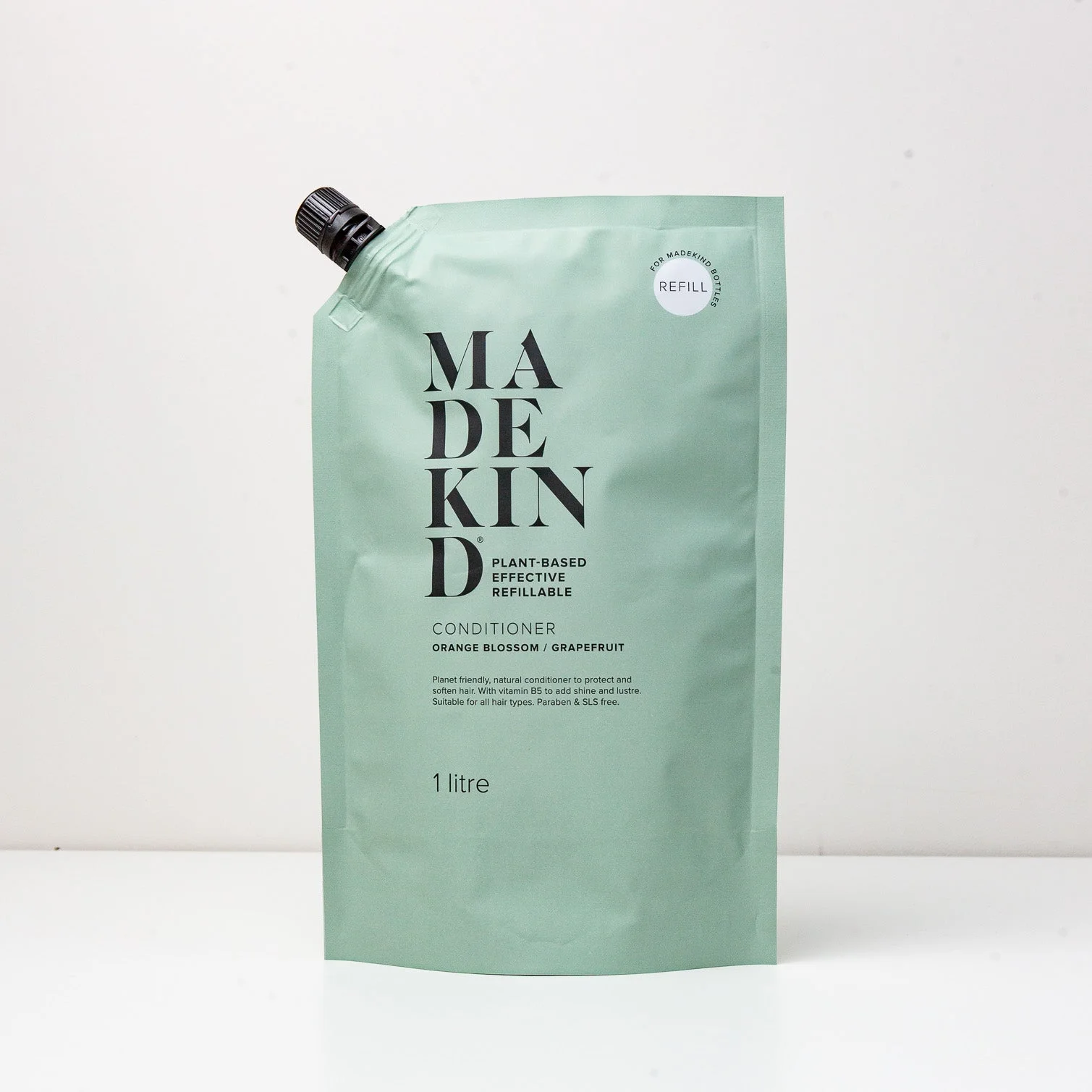 A green pouch labeled 'MADEKIND' containing hair conditioner. The packaging indicates it is a 1 litre refill of conditioner with orange blossom and grapefruit scents.